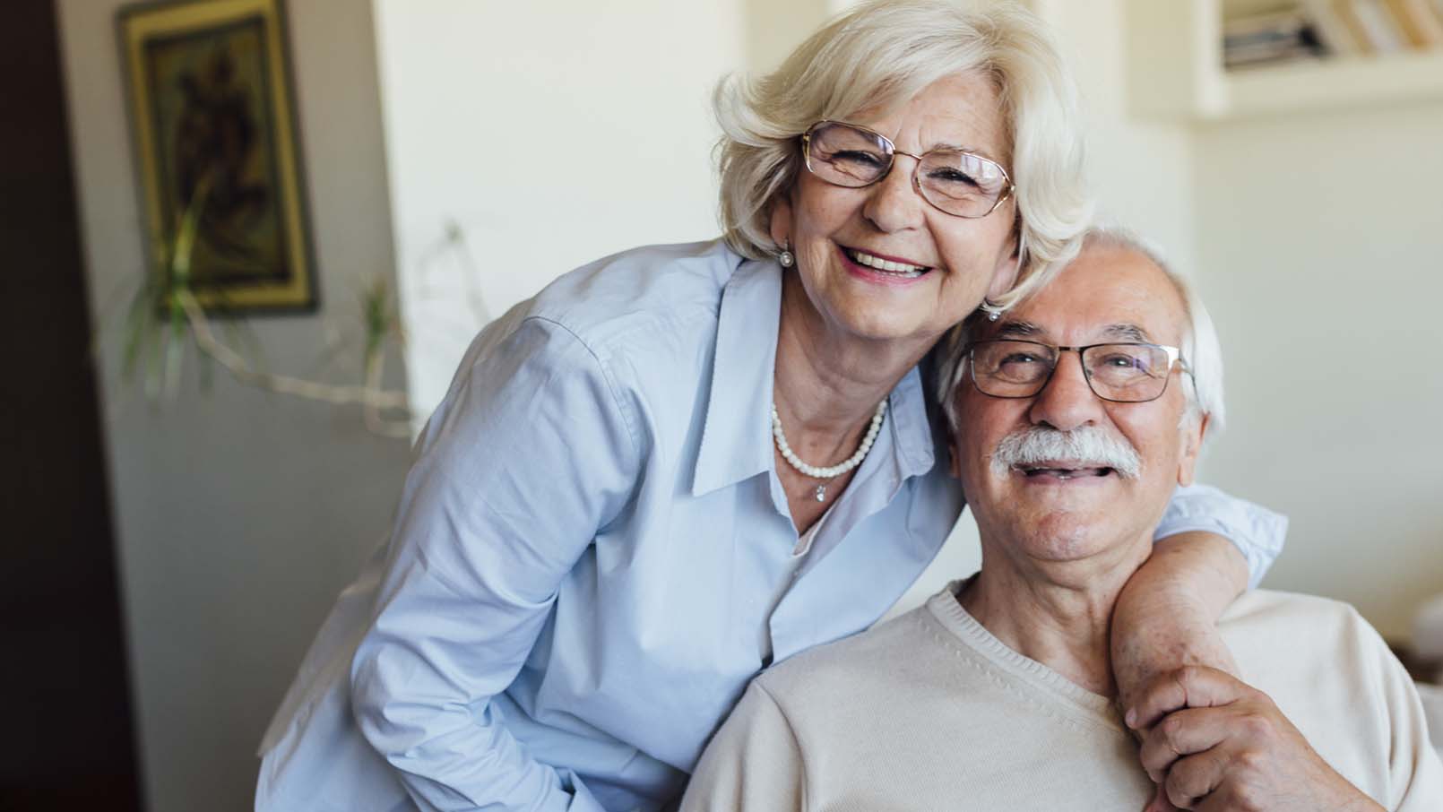 A smiling elderly white couple sit together inside of their Erickson Senior Living apartment home.