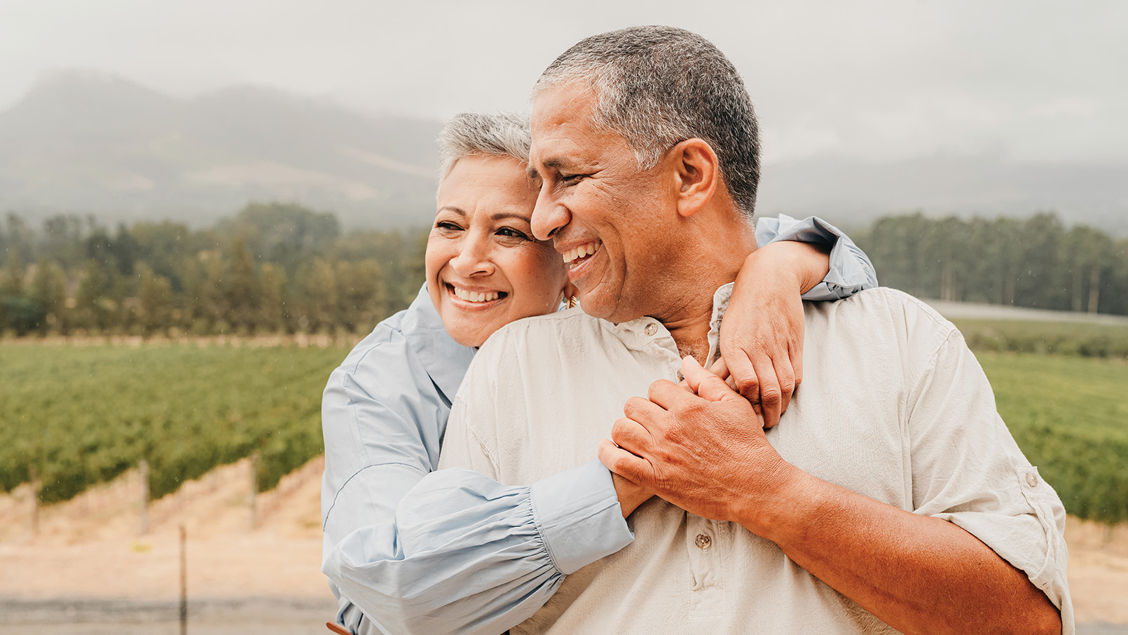 Older Hispanic couple, with the woman hugging the man, standing in front of a scenic outdoor backdrop with mountains and a vineyard.